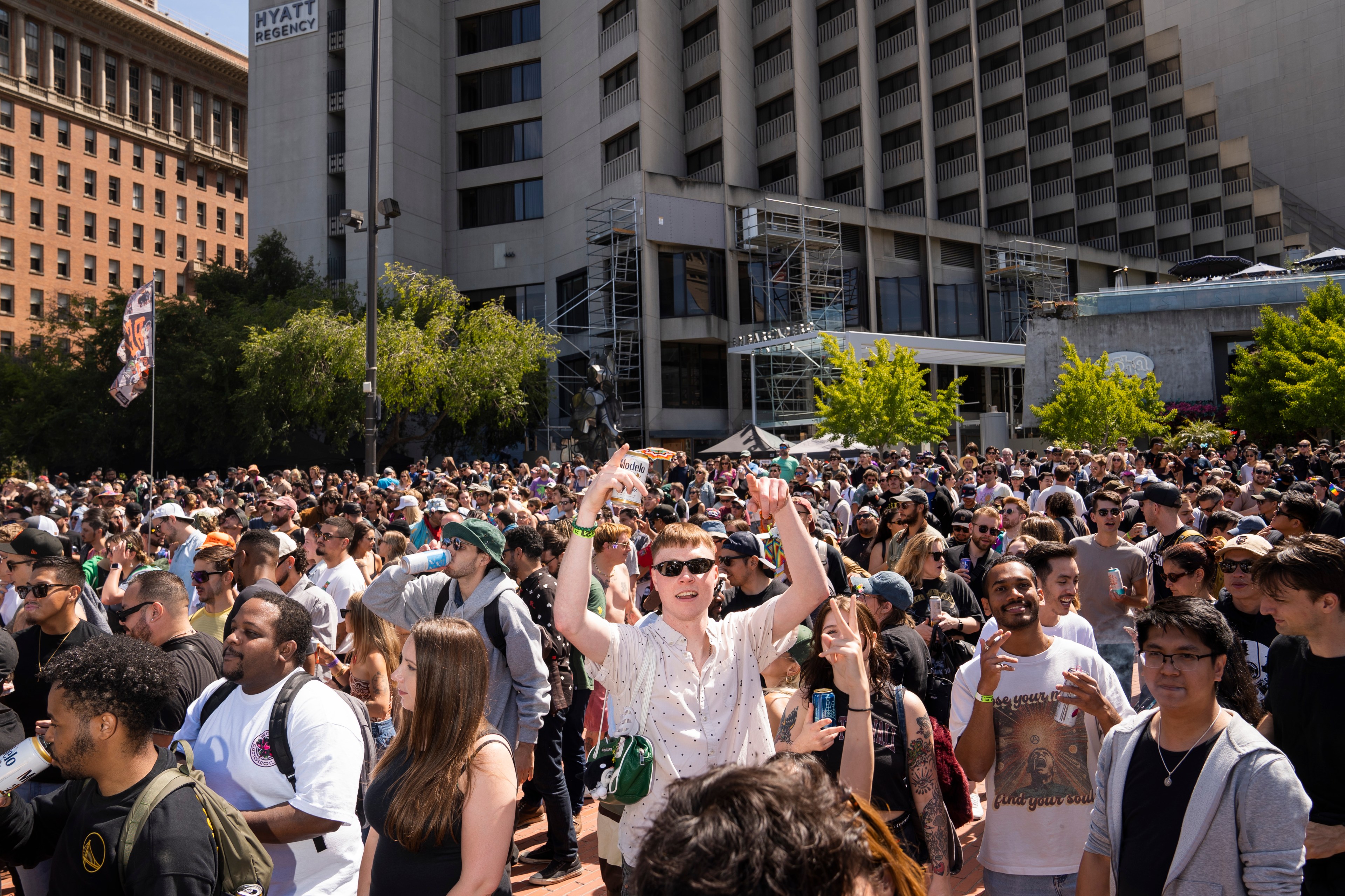 A large crowd of people is gathered outdoors in front of tall buildings. Many individuals are smiling, wearing sunglasses, and holding drinks under a sunny sky.