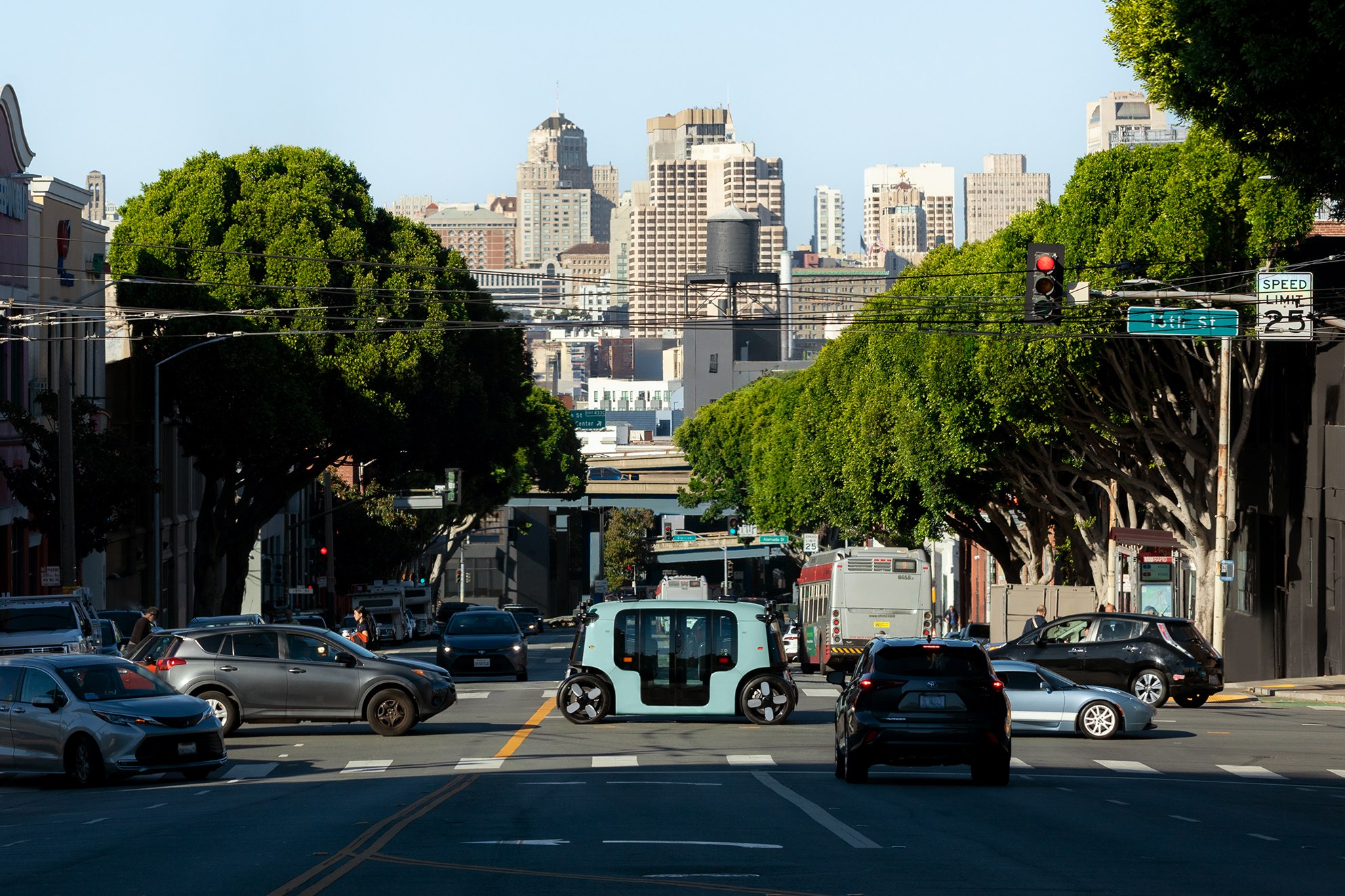 A busy city street features cars and a unique small vehicle in the center. Lush green trees line both sides, with tall buildings rising in the background.